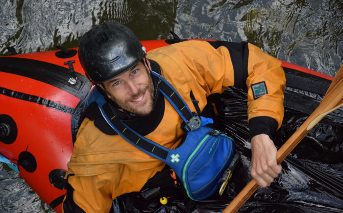Man in a red kayak with a paddle, blue life vest, orange top and black helment looks up from the water.