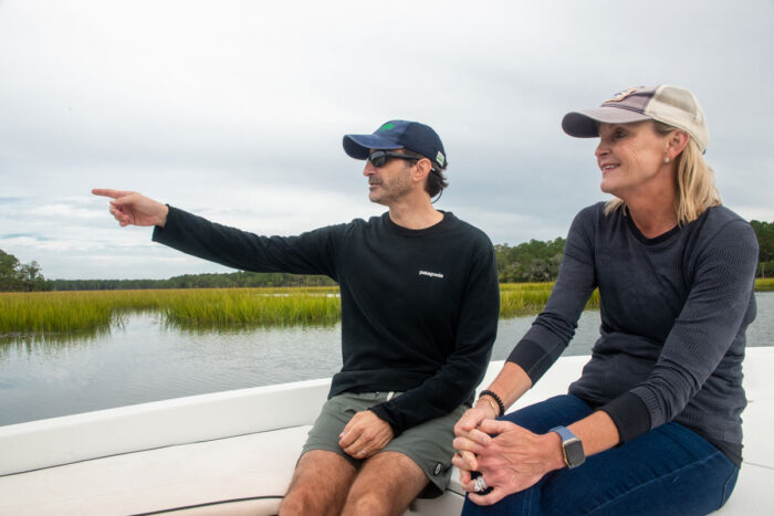 A man and woman sit on the front of a small power boat looking out with water and wetlands in the background.