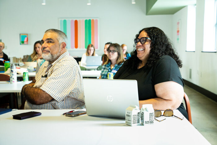 A man and woman are sitting in the front a classroom-like setting, with some other people behind them. She has a laptop and a big smile.