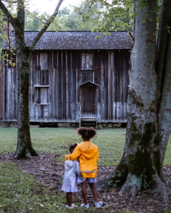 Two young girls stand with their arms around each other looking at an old gray boarded up building.