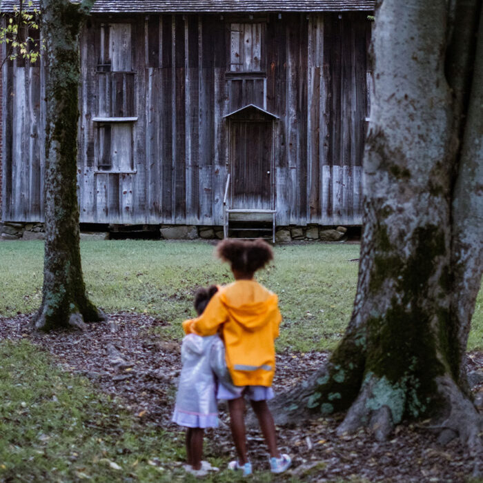 Two young girls stand with their arms around each other looking at an old gray boarded up building.
