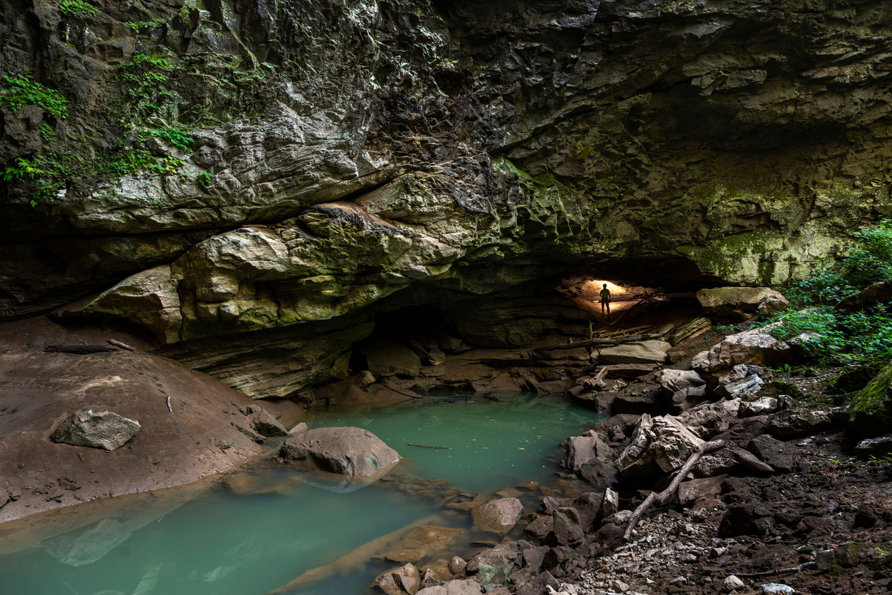 A stream surrounded by rock leads into a rock wall where a small glow of light surrounds someone entering into a cave you can't see.