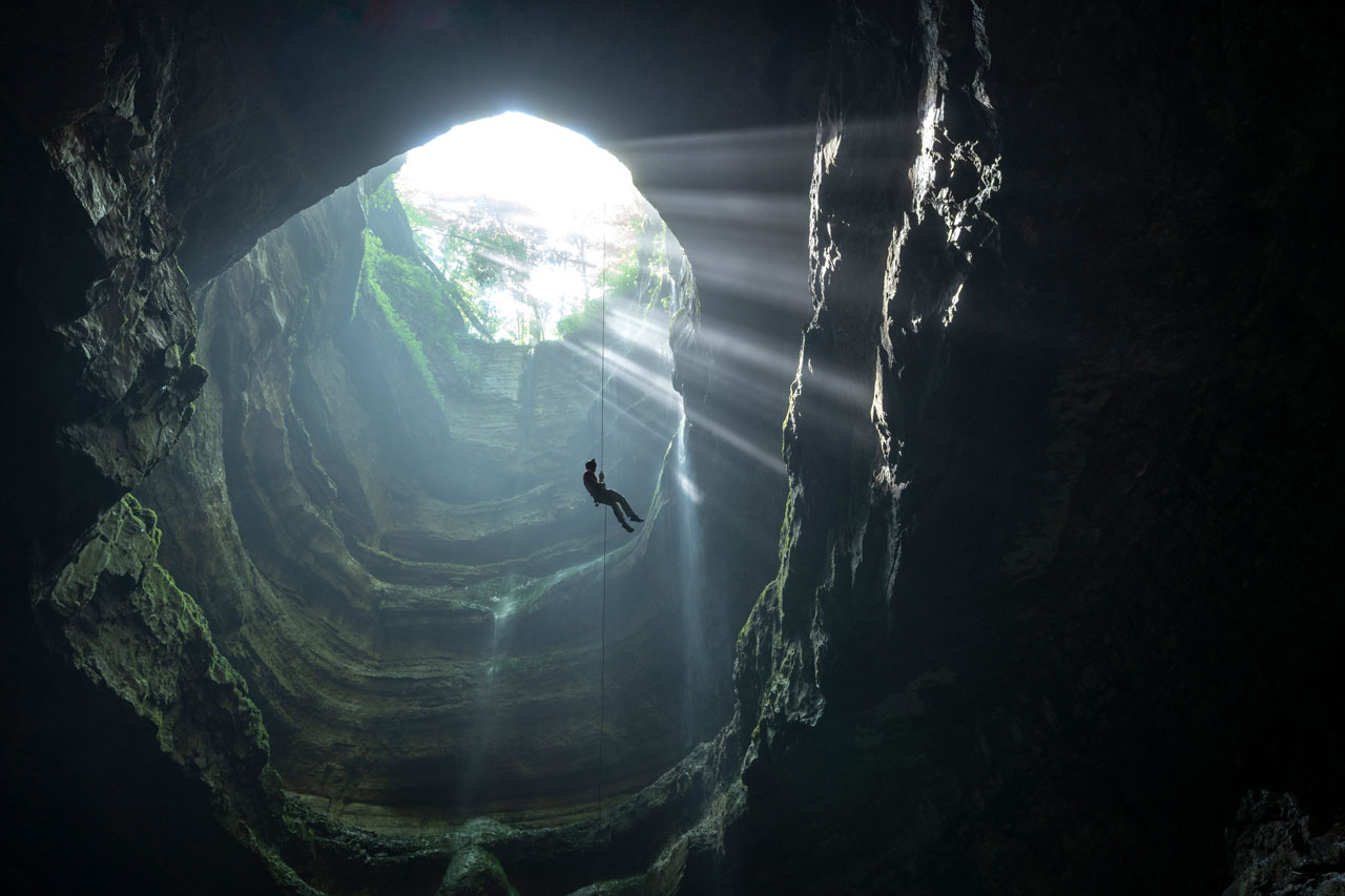 Looking up at someone belaying down a rope through a large underground shaft lit by rays of light.