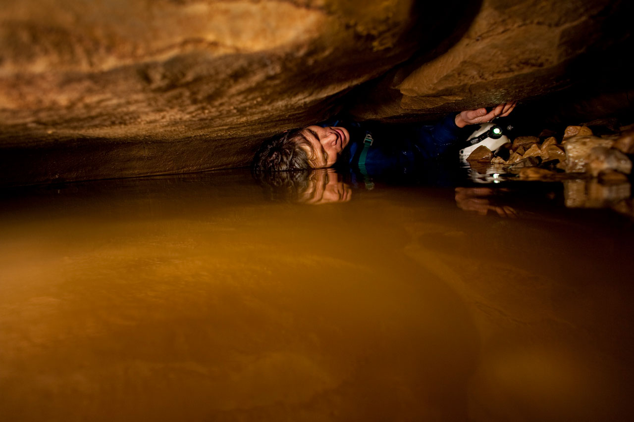 A man wearing a climbing helmet and headlamp squeezes through a narrow space between two huge rocks.
