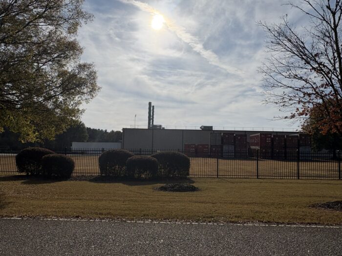 A wide shot of a beige, rectangular industrial building under a bright, hazy sun. The facility is enclosed by a black metal fence and features two tall, thin exhaust stacks rising from the roof. To the right of the building, several shipping containers are stacked in a yard.