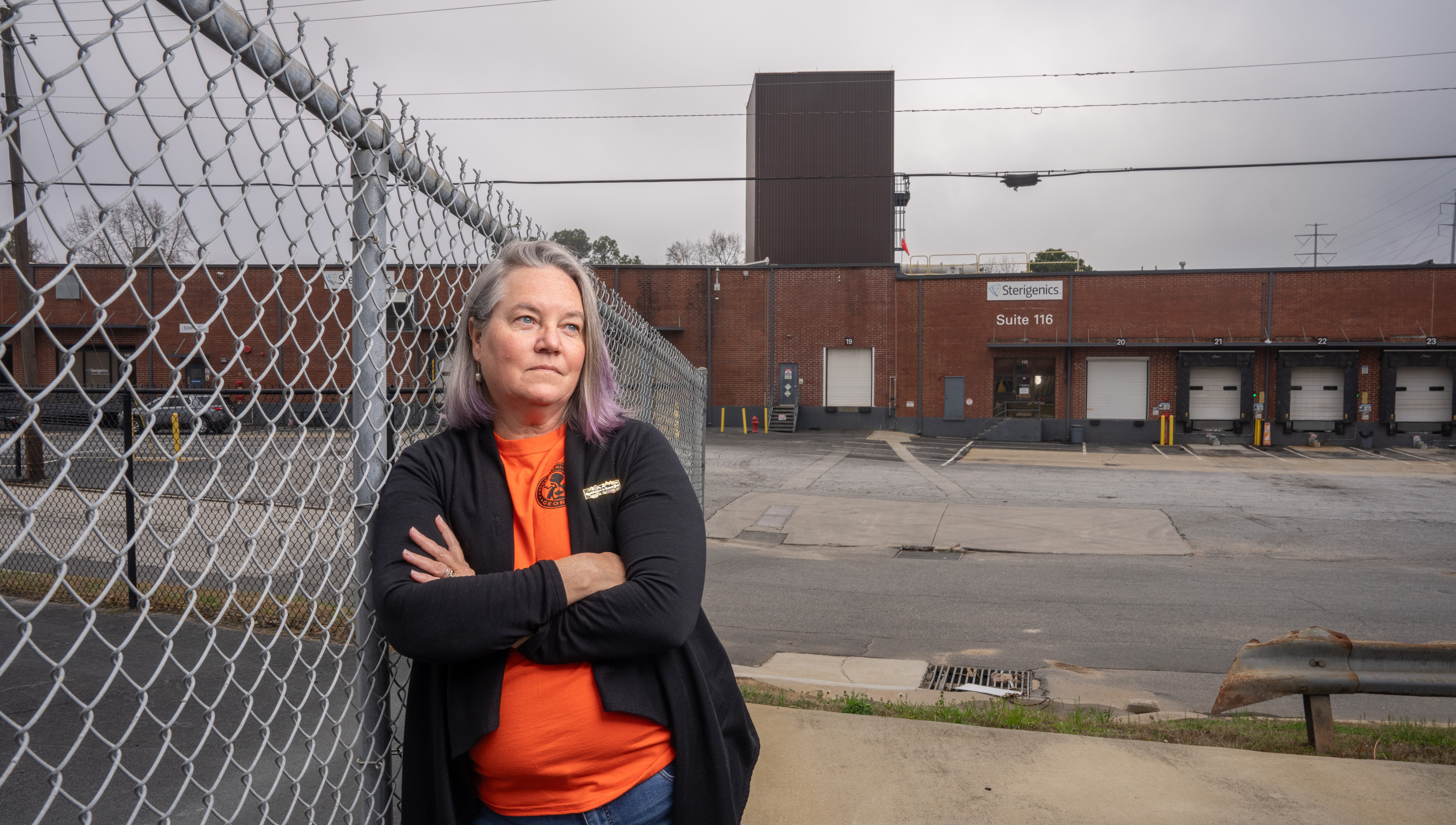 A white woman with greying hair in an orange tshirt and fleece jacket leans against a chainlink fence with a drab brick industrial building behind her.