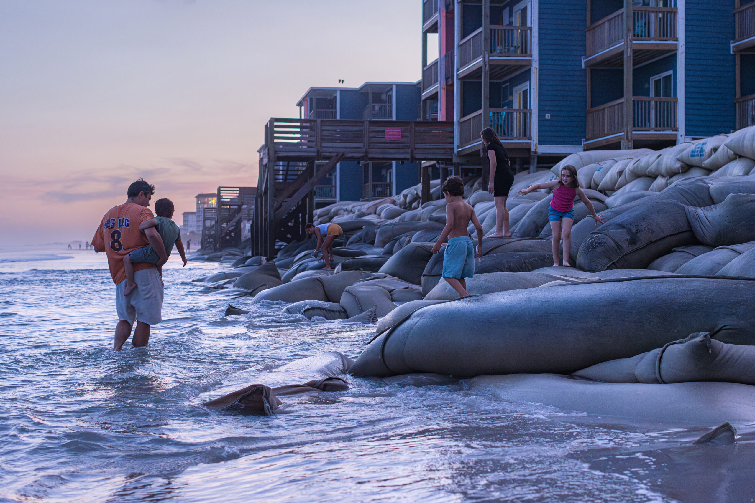 A dad walks down the beach carrying a kid and watches another playing on the sand bags piled up in front of beach front homes.