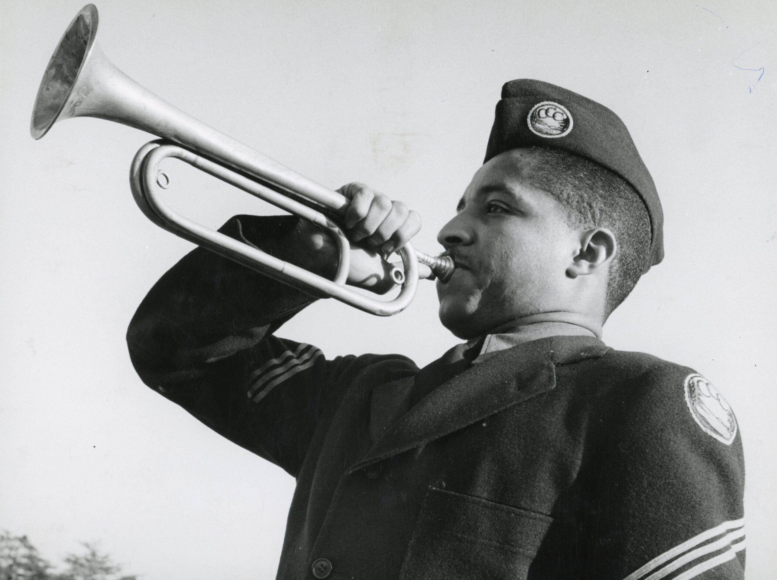 A black and white photo of a Black man in a uniform blowing a bugle.