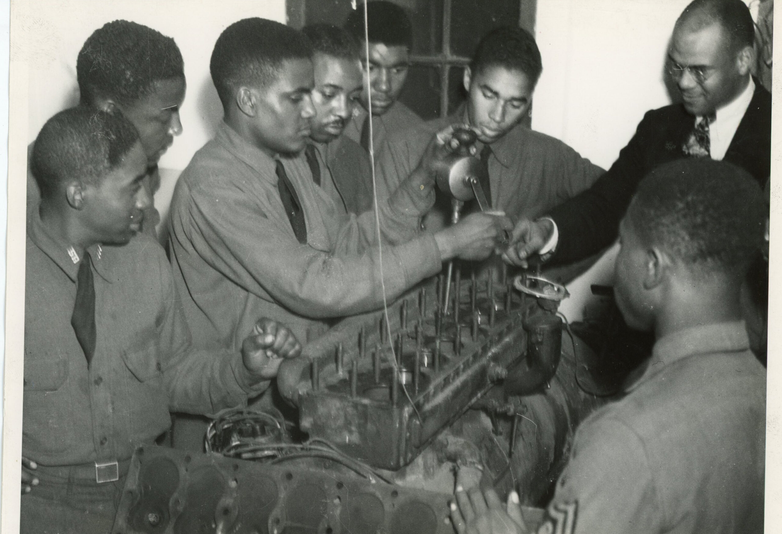 A group of Black young men in uniforms lean over to learn more about an engine.