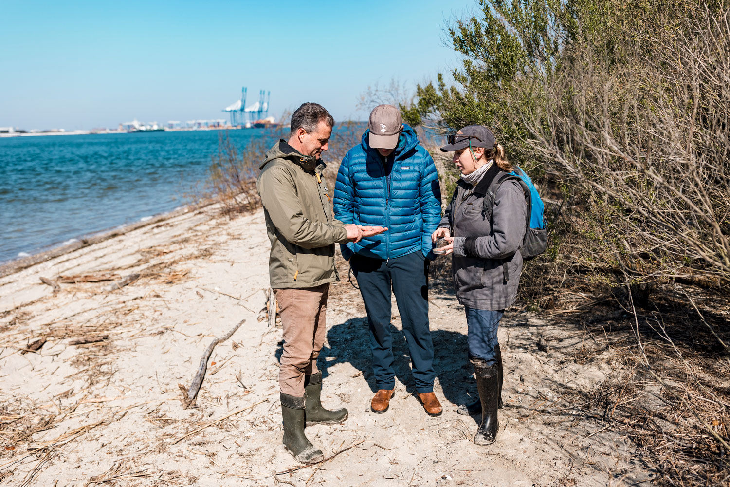 Three people stand on a beach in warm clothes looking at something small in one persons hand. Two port cranes are visible across the water.